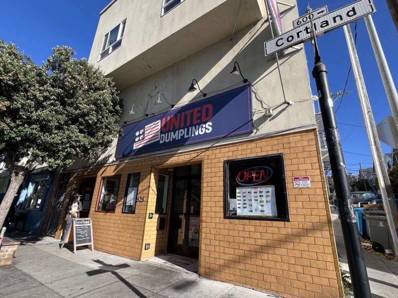 Street view of United Dumplings restaurant with a large sign above the entrance. Sidewalk chalkboard menu and "OPEN" neon sign in the window. Nearby intersection sign shows Cortland Ave and 60th St.