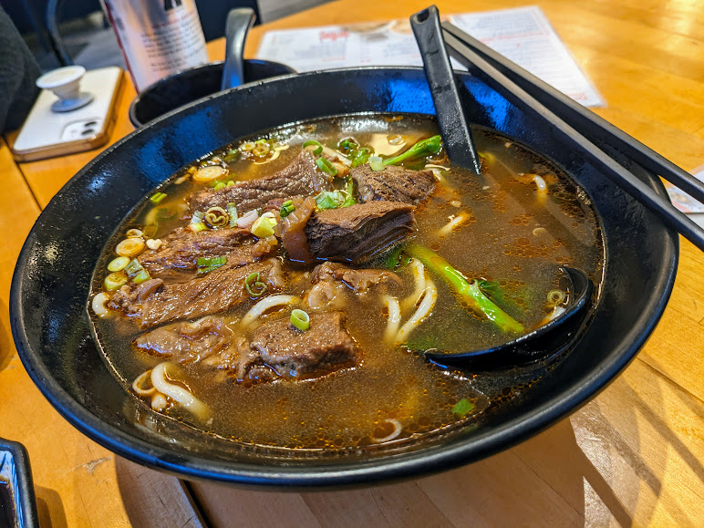 A bowl of beef noodle soup with chunks of beef, green onions, and noodles in a dark broth, served with black chopsticks and a spoon on a light wooden table.