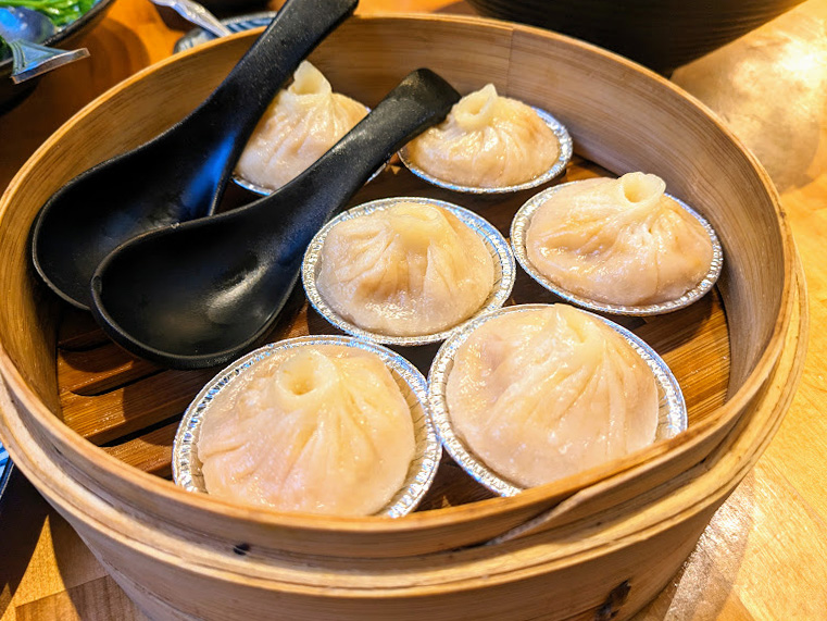 A bamboo steamer with six steamed dumplings in metal cups and two black spoons inside.