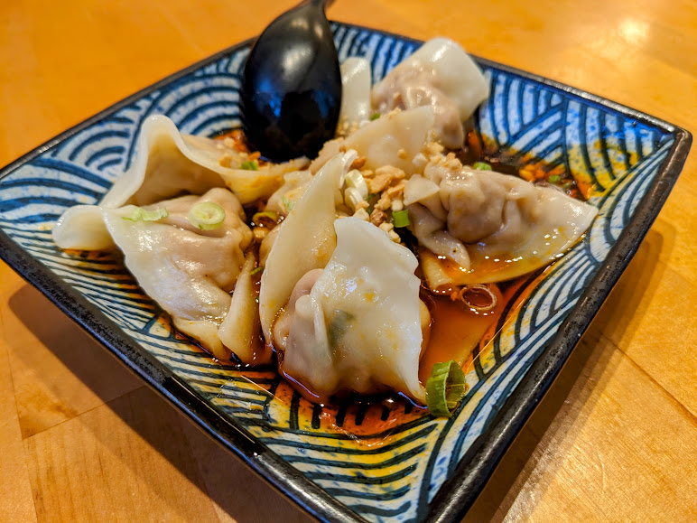 A square plate of dumplings topped with chopped green onions and submerged in a sauce, with a black spoon on the side.