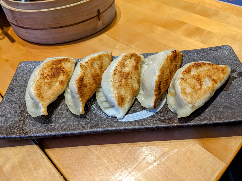 A rectangular plate with five pan-fried dumplings arranged in a row is placed on a wooden table. A bamboo steamer is visible in the background.