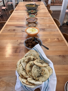 A variety of Mediterranean dishes, including pita bread, falafel, hummus, and other dips, are arranged in bowls along a long wooden table.