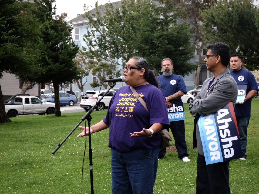 A person speaks into a microphone at an outdoor event while others stand nearby holding signs that read "Ahsha Mayor.