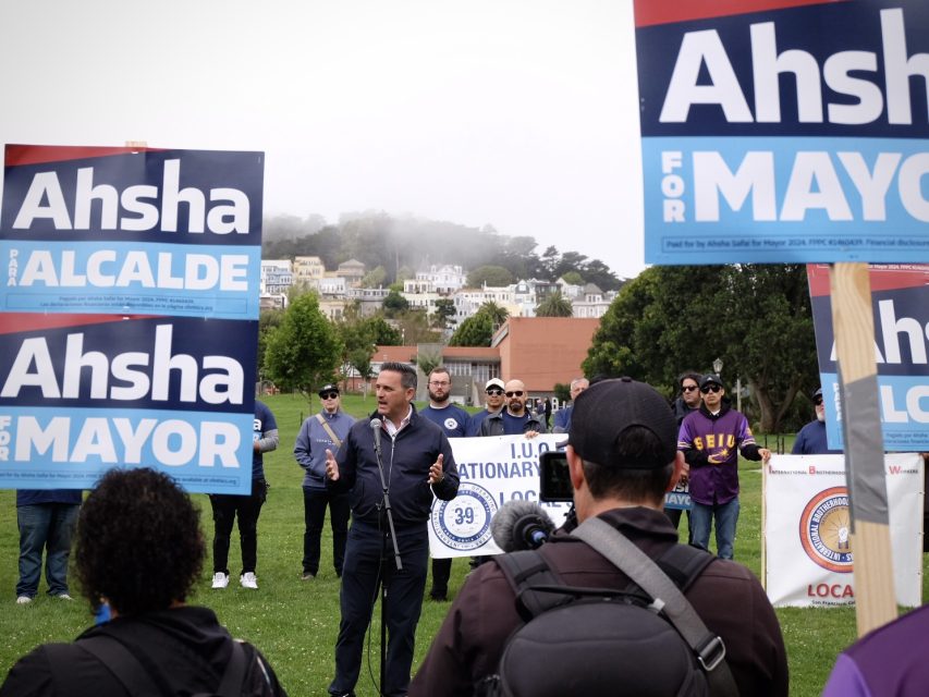 A speaker addresses a crowd at an outdoor event, surrounded by people holding "Ahsha for Mayor" signs. Some attendees wear union-related shirts and buttons. Houses and greenery are visible in the background.