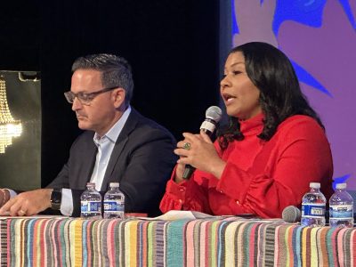A man in a suit and a woman in a red blouse sit at a table with microphones and bottled water in front of them. The woman is speaking into a microphone.