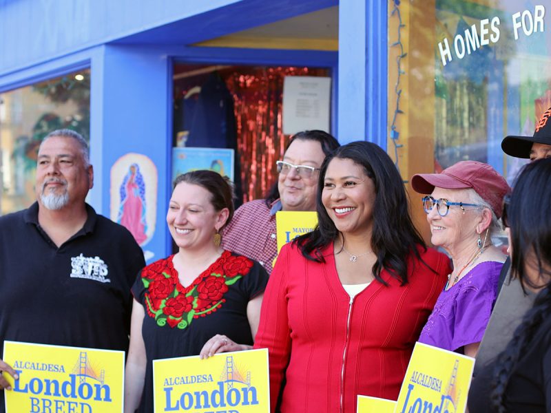 A group of people stands outside a blue-painted building, holding yellow signs that say "London Breed." One woman in a red top is centered. Some are smiling, and the setting appears to be a campaign event.