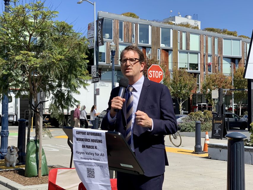 A man in a suit speaks into a microphone at an outdoor event. A sign on the podium reads, "Support Affordable Workforce Housing on Parcel K, Hayes Valley For All." Modern buildings are in the background.