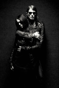 Two people dressed in black leather jackets stand in a dark setting, with one embracing the other from behind.