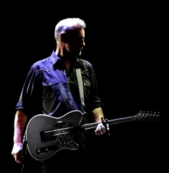 A person stands in a dark setting, holding a guitar and looking to the side while wearing a dark shirt.