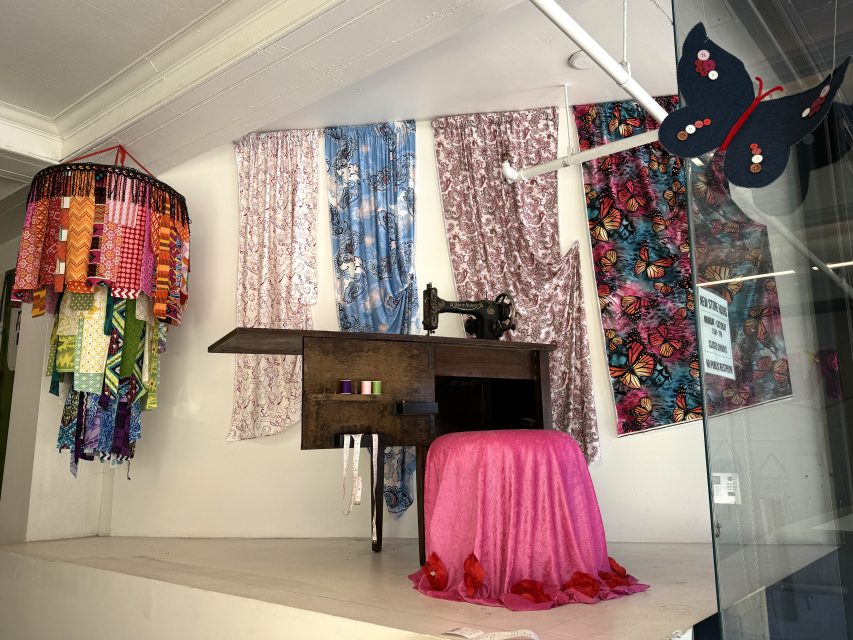 A display features an antique sewing machine on a wooden table with a pink draped stool, colorful hanging fabric samples, and butterfly-themed decorations in the background.
