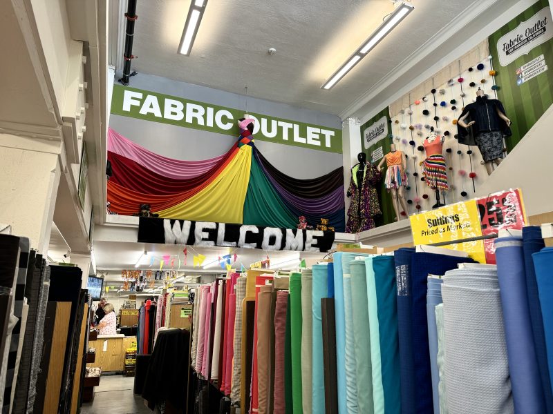 Interior of a fabric outlet store with numerous rolls of fabric on display. A colorful fabric arrangement and a "Welcome" banner hang above. Clothing and signage are visible on the walls.