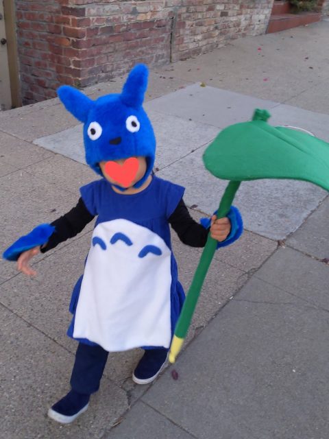A child in a blue and white costume with a large green prop stands on a sidewalk near a brick wall.