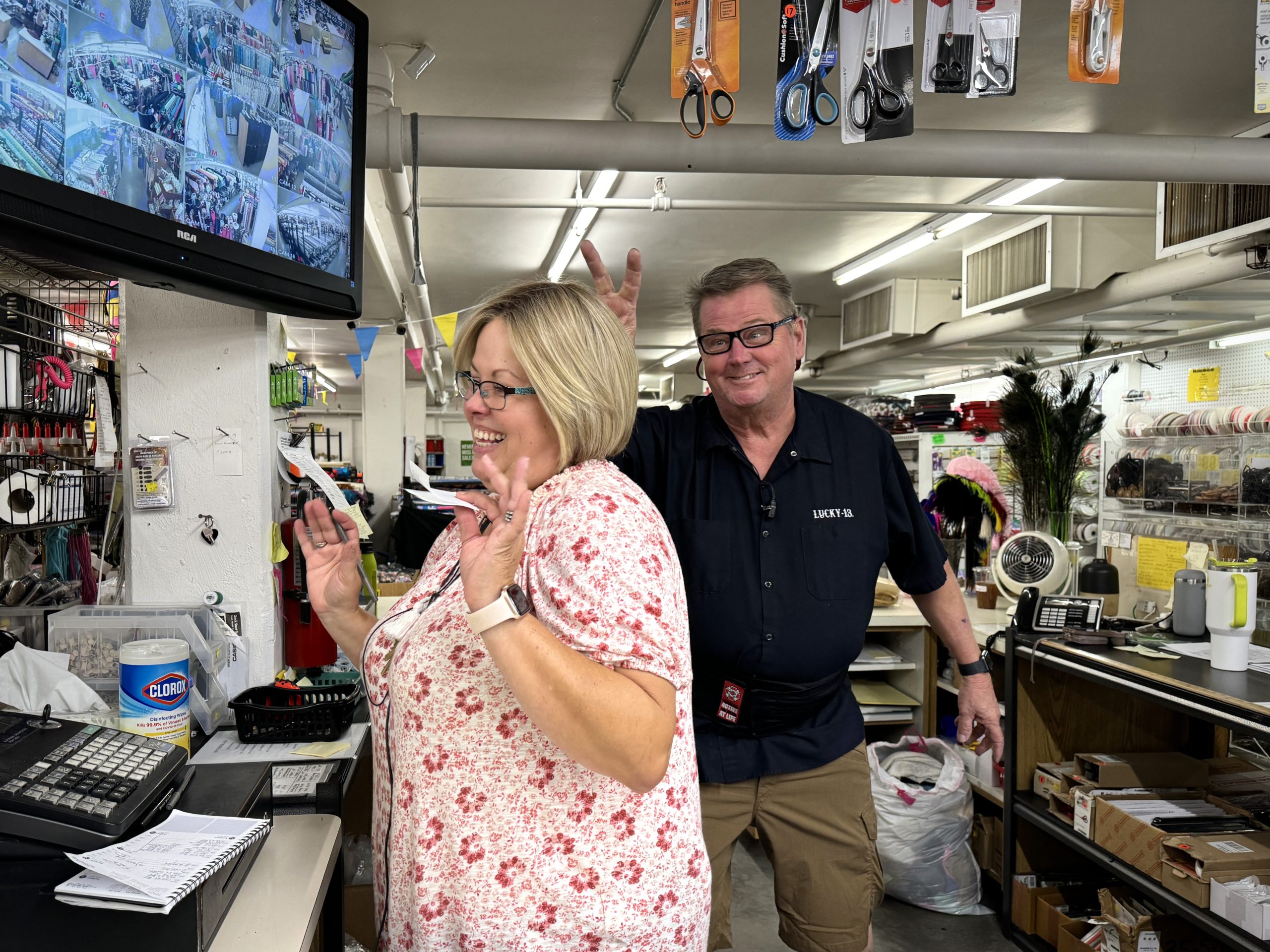 Two people in a store: a woman smiles while making a peace sign on one hand, and a man behind her makes a playful gesture with his fingers. They stand near a counter with various items and a monitor.