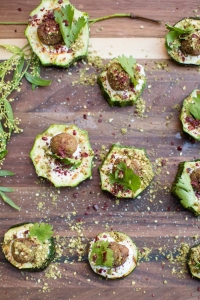 A wooden board with several zucchini slices topped with a dollop of white sauce, sprinkled with green herbs and red spices, garnished with small meatballs and cilantro leaves.