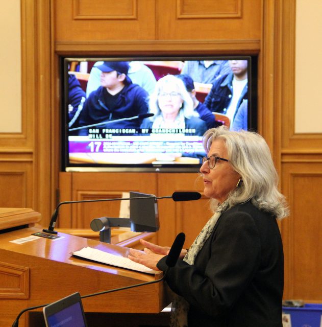 An individual speaks at a podium with a microphone in an uptown conference room, while a screen behind shows another person and displays text.