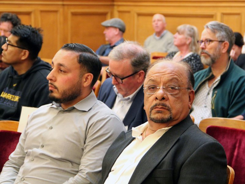 A group of people sits attentively in a wood-paneled room in the heart of uptown. Some are focused on the front, while others appear to be deep in thought.
