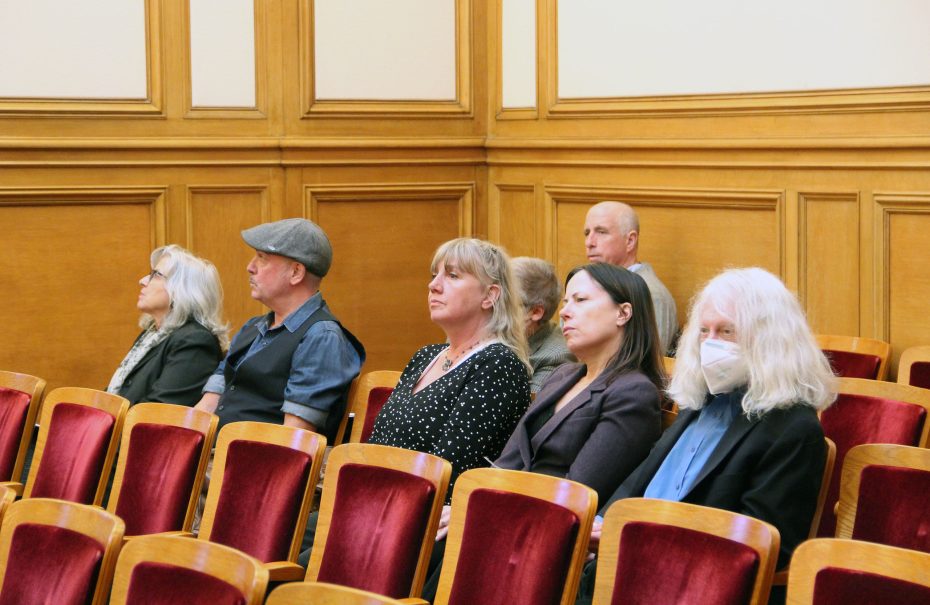 Six people are seated in an uptown room with wooden paneling and red velvet chairs, attentively watching something ahead. One person is wearing a mask.