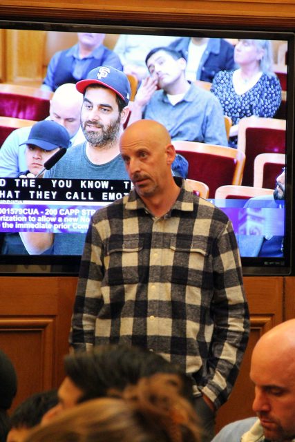 A bald man in a checkered shirt speaks in front of a seated audience. Meanwhile, on the screen in the background, someone sporting an uptown look with a logo hat listens intently.