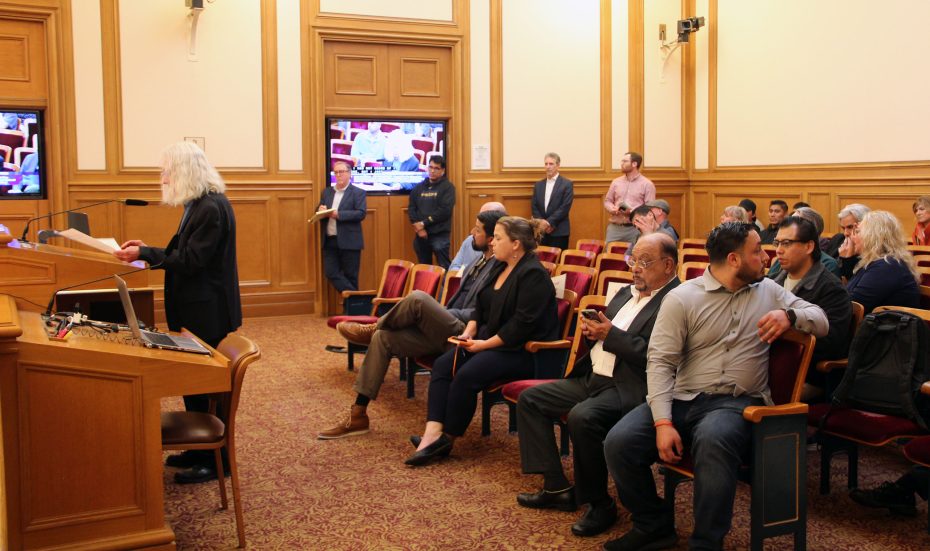 A group of people seated and standing in a room with wooden paneling, some attentively watching and others on their devices, while a person speaks at a podium in an elegant uptown setting.