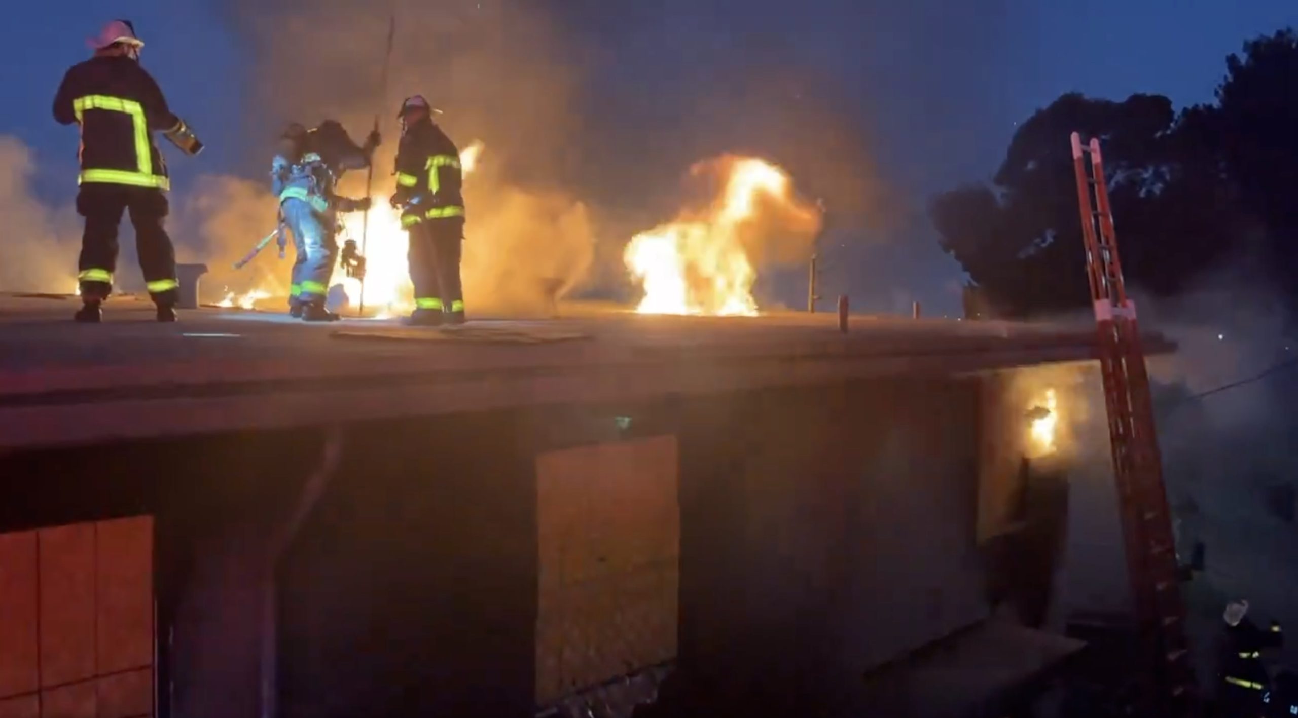 Firefighters in protective gear work to extinguish a fire on the roof of a building at night; flames and smoke are visible. A ladder is propped against the building.