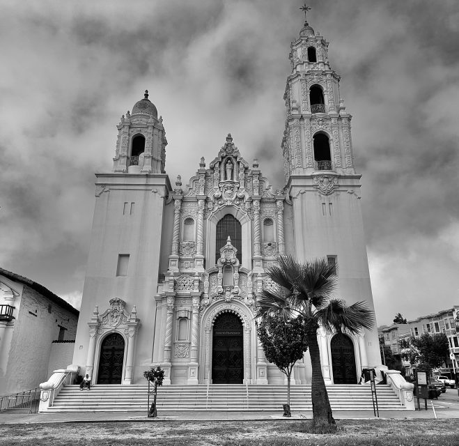 Black and white image of Mission Dolores basilica with twin towers, detailed facade, and multiple arched entrance doors. A lone palm tree stands in front of the church.
