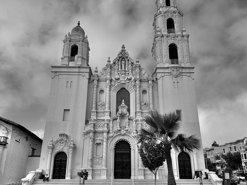 Black and white image of Mission Dolores basilica with twin towers, detailed facade, and multiple arched entrance doors. A lone palm tree stands in front of the church.