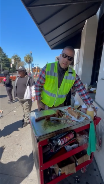A street vendor in a reflective vest stands by a food cart with condiments and prepared food on a sunny day. People are visible in the background.