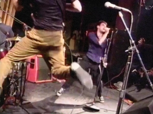 Musician jumping in mid-air while playing guitar on stage; vocalist singing into a microphone; various instruments and equipment visible, including drums and amplifiers.