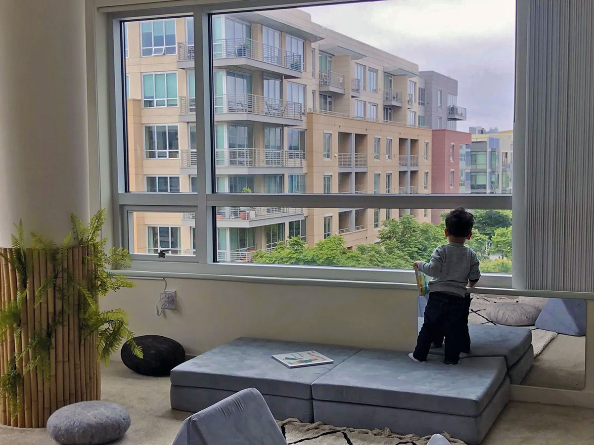 A toddler stands on cushions, peering out a large window at the modern Lai apartment complex. Nearby plants and decor add a touch of warmth in the foreground.