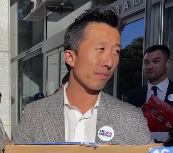 A man wearing a grey blazer and "SF DEMS" pin stands outdoors, holding a box, while Lai, in a navy suit, is partially visible in the background with a gift bag.