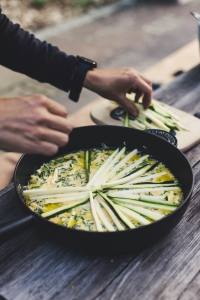 A person arranges zucchini slices on top of a dish in a skillet on a wooden table.