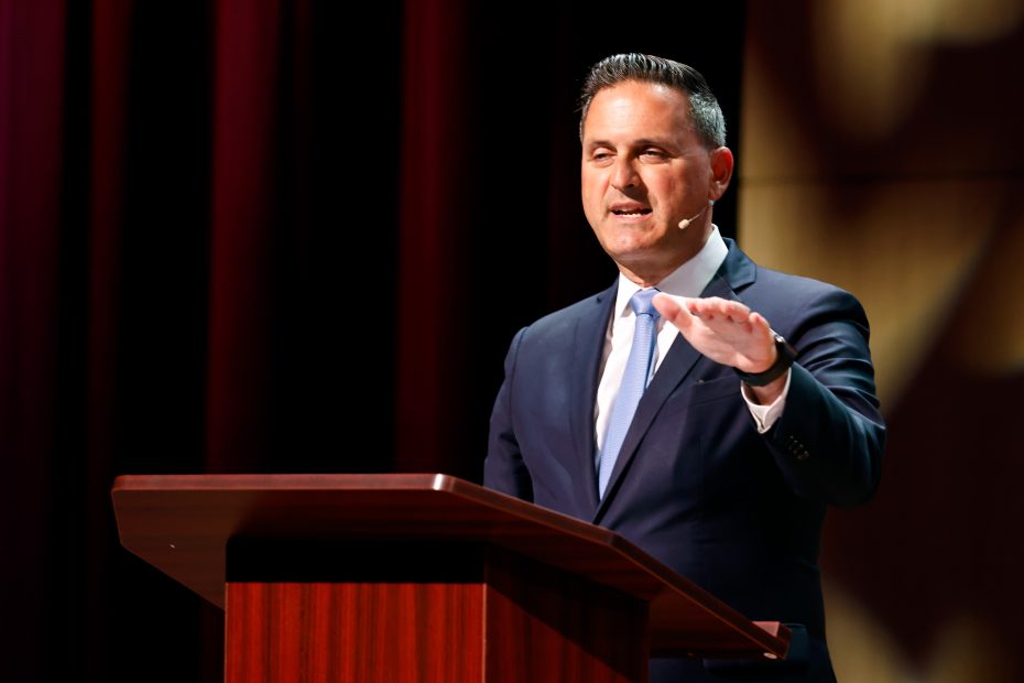 A man in a suit speaks at a wooden podium on a stage, with a red curtain in the background. He is gesturing with his right hand while speaking into a headset microphone.