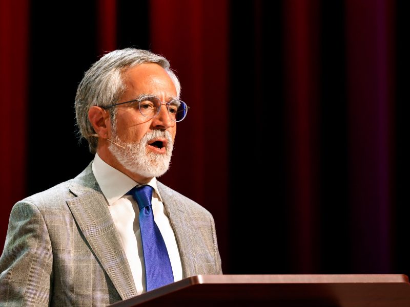 A man with grey hair and beard, wearing glasses, a grey suit, white shirt, and blue tie, speaks at a podium with a maroon curtain in the background.