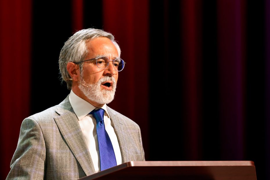 A man with grey hair and beard, wearing glasses, a grey suit, white shirt, and blue tie, speaks at a podium with a maroon curtain in the background.