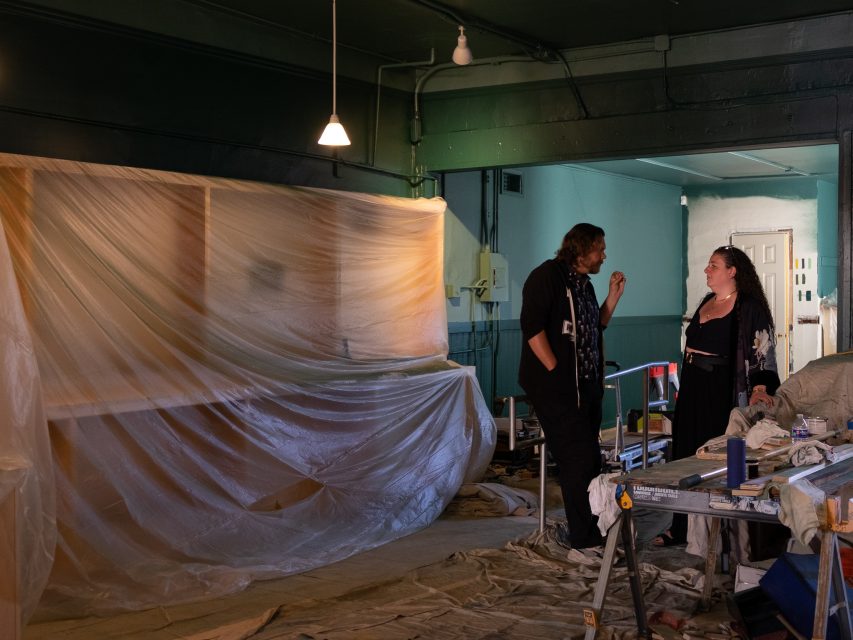 Two people engage in conversation inside a room under renovation, with plastic sheeting draped over furniture and various tools scattered on the floor.