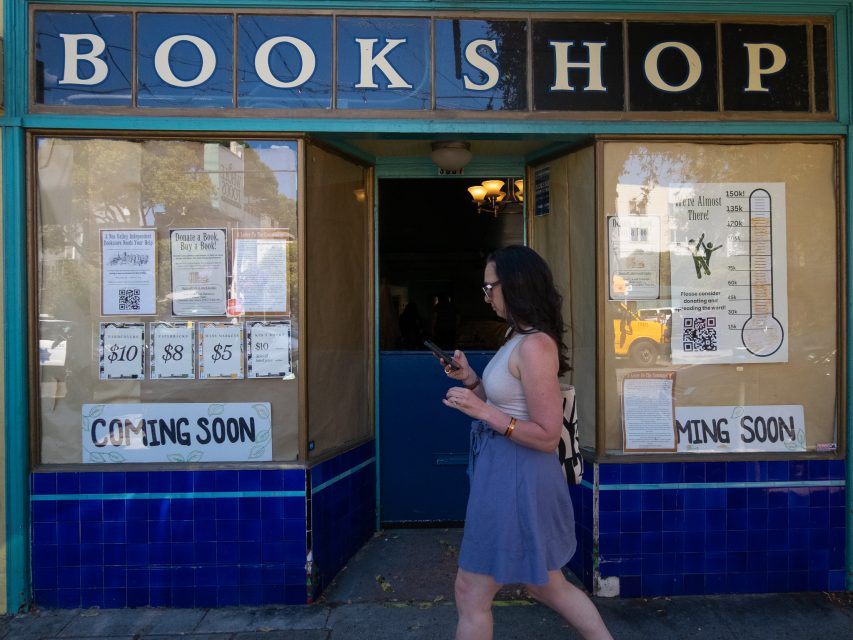 A woman walks past a storefront with a "BOOKSHOP" sign above and partially covered "COMING SOON" signs on the windows.
