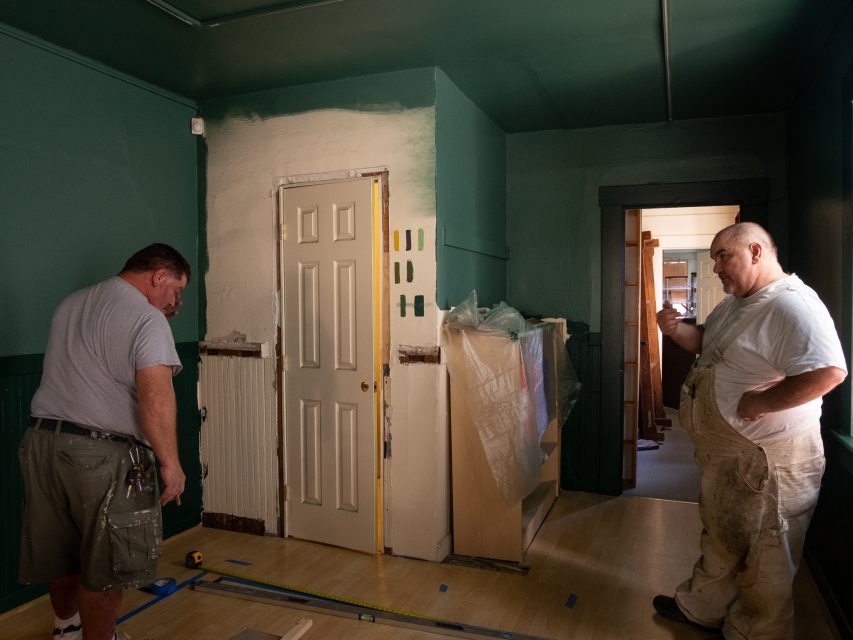 Two men are discussing while renovating a room. Walls are painted green, and partially painted samples are near a door. Various tools and materials are on the floor.