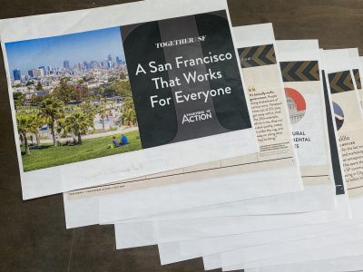 Printed documents are spread out on a table, with the top sheet displaying a cover page titled "A San Francisco That Works For Everyone" alongside an image of a park and the city skyline.