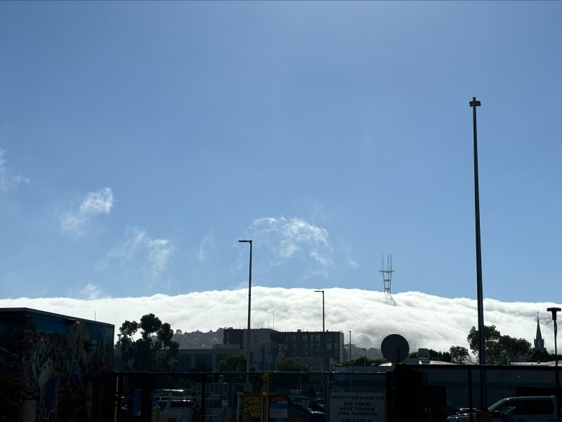 A dense layer of fog blankets a cityscape under a bright blue sky, with tall utility poles and a radio tower rising above the fog.