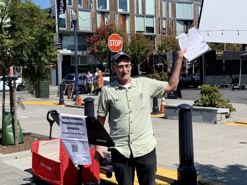 A man stands at a street corner holding up papers, next to a sign promoting affordable housing. A stop sign and several buildings are visible in the background.