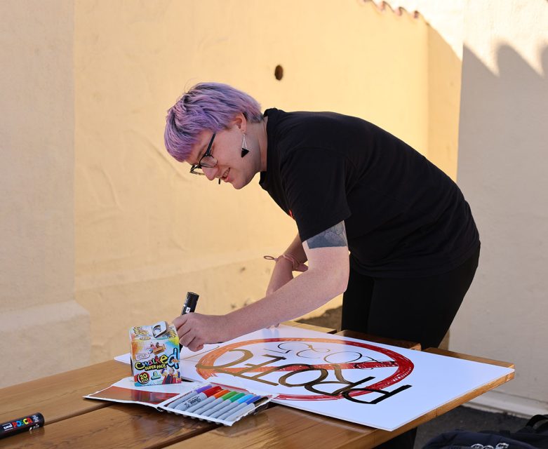 Person with short, purple hair and black clothing is standing at a table and drawing on a white poster with colorful markers.