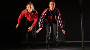 Two people in casual athletic wear are captured mid-jump against a dark background. Both are looking forward and appear to be in motion.