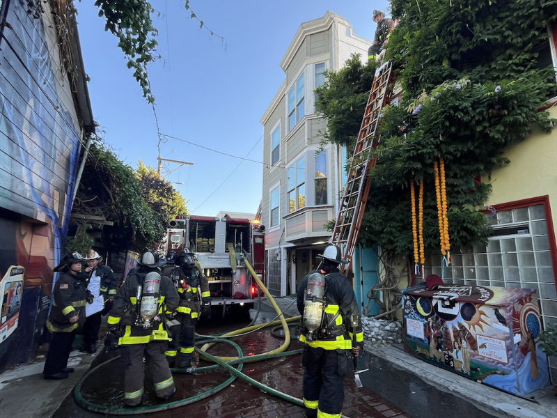 Firefighters work at the scene of a building with a fire truck parked nearby. Some are on the ground and others are climbing ladders. The area is surrounded by colorful murals.