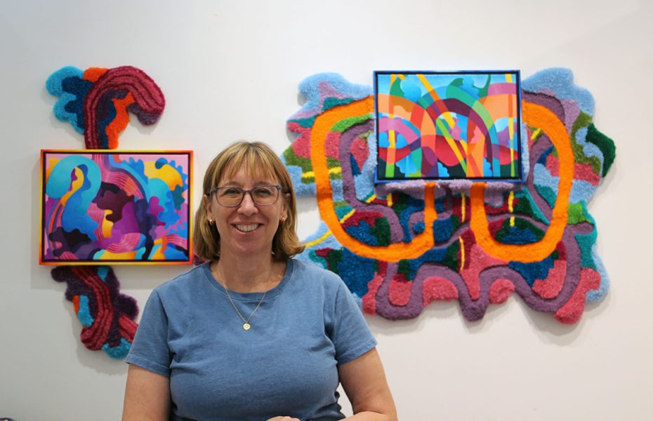 A woman in a blue shirt stands smiling in front of a white wall with colorful abstract artwork displayed.