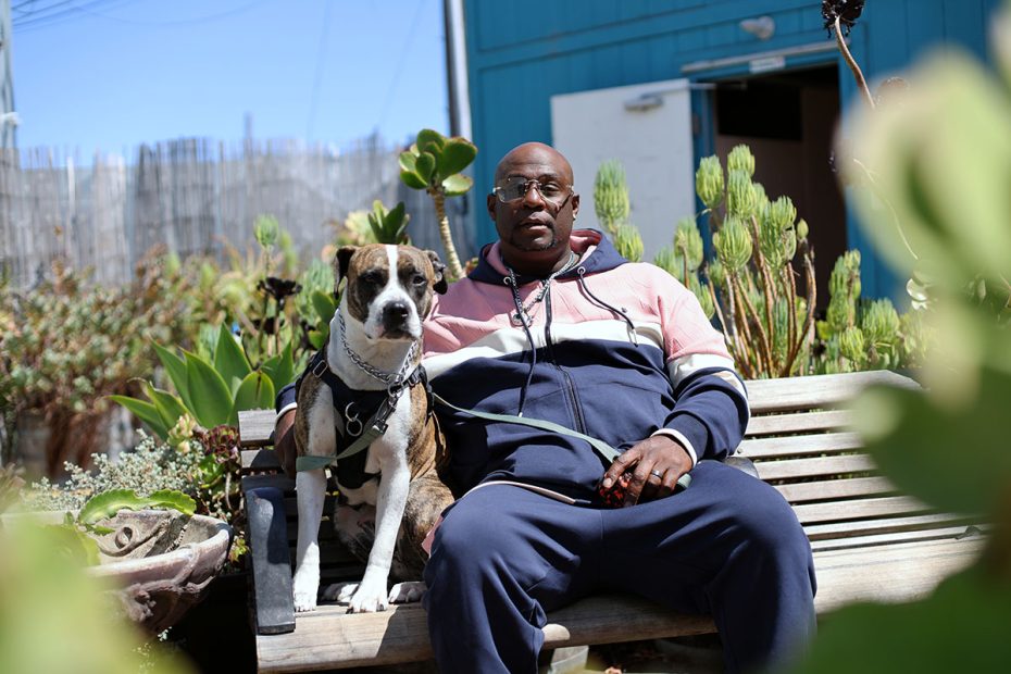 A man in a tracksuit sits on a bench outside, holding a dog on a leash. The setting includes plants and a blue building in the background.