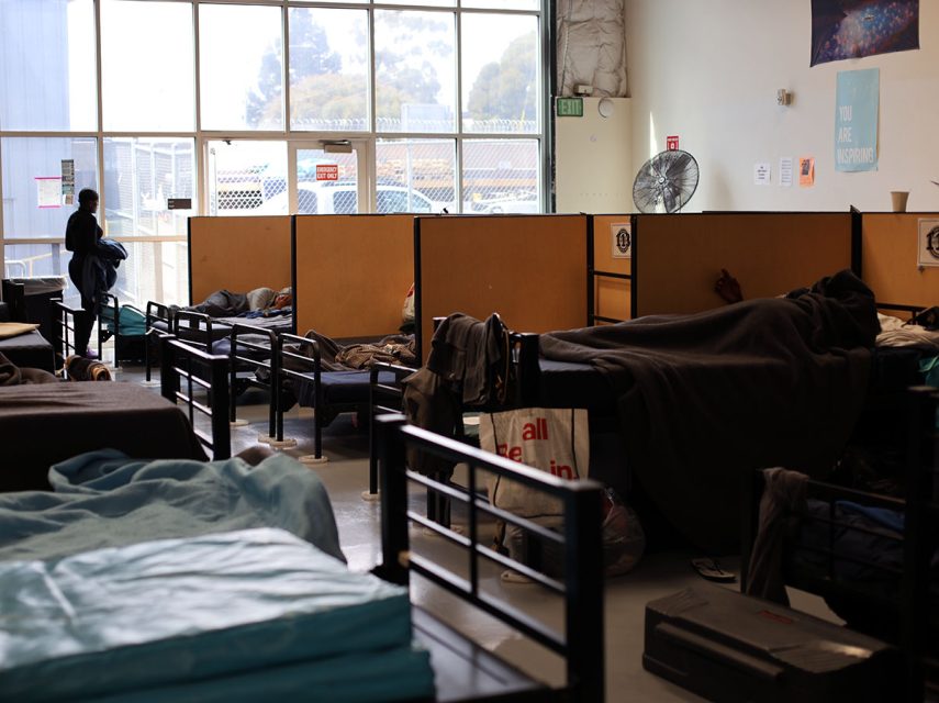 A homeless shelter with occupied beds and personal belongings; a person stands near a window.