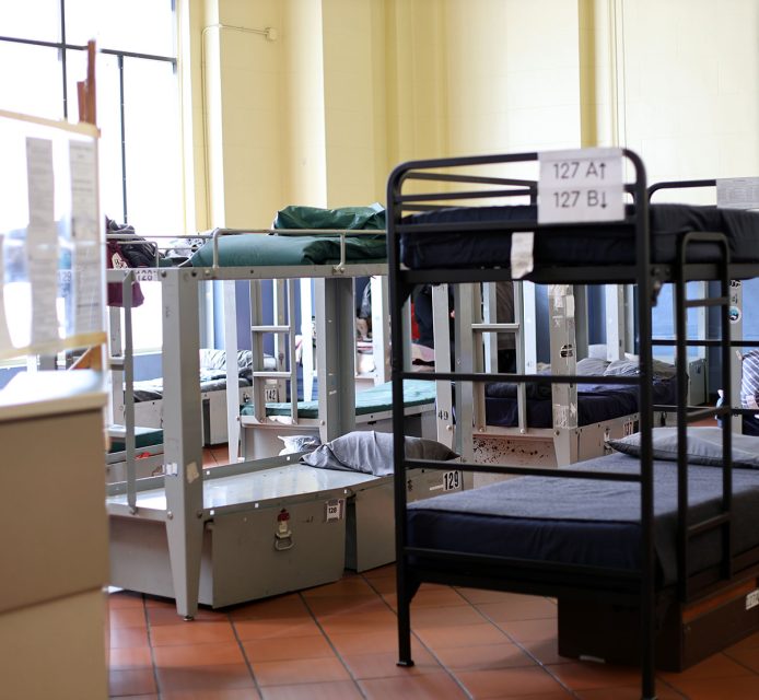 A room with multiple metal bunk beds, some unmade, and personal belongings. The walls are light yellow, and the floor is tiled. There are windows providing natural light.