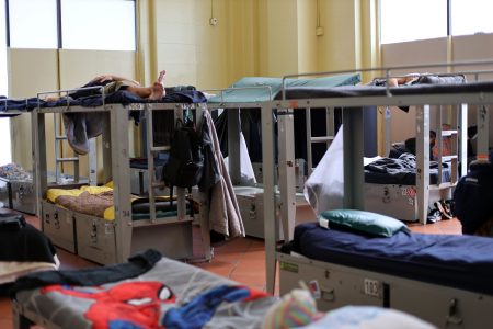 A room with multiple metal bunk beds, some occupied. Bedding, personal belongings, and hanging clothes are visible. The space is well-lit by large windows.
