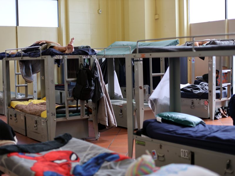 A room with multiple metal bunk beds, some occupied. Bedding, personal belongings, and hanging clothes are visible. The space is well-lit by large windows.
