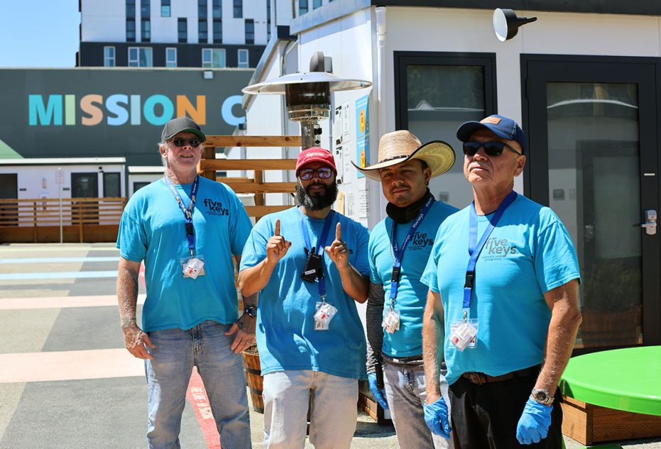Four men wearing blue "Five Keys" shirts and ID lanyards stand in an outdoor area with a building and a sign reading "MISSION" in the background.
